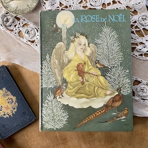 Peut inclure: Livre vintage intitulé "La Rose de Noël" avec une couverture verte représentant une illustration d'ange jouant du violon, entouré d'oiseaux et d'une scène de forêt enneigée. Le livre est sur une surface en bois.