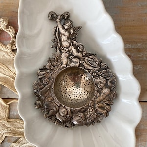 May include: A silver-toned metal tea strainer featuring an ornate relief design of cherubs, flowers, and fruit surrounding a central perforated bowl. The handle is shaped like a cherub figure, and the entire piece has a dark, aged patina finish.