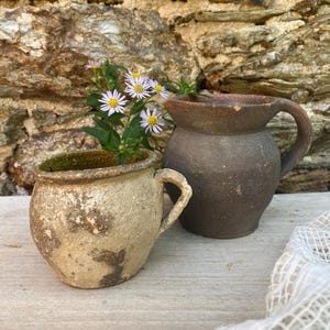 Op de afbeelding: Een crèmekleurige keramische pot met een handvat en een donkerbruine keramische kan met een handvat, beide met een textuur. Een kleine groene plant met lichtpaarse bloemen met gele harten staat in de crèmekleurige pot.