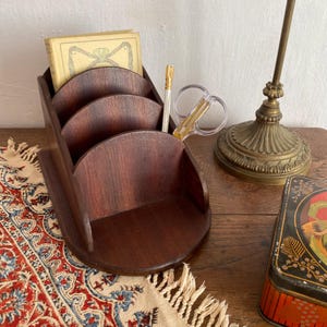 May include: A dark brown wooden desk organizer with three curved compartments. A small book, a gold-tipped pen, and clear scissors are in the organizer. A vintage lamp and a decorative tin are also visible.