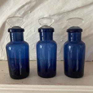 Op de afbeelding: Drie kobaltblauwe glazen apothekersflessen met doorzichtige glazen stoppers. De flessen hebben een afgeronde vorm en worden op een wit oppervlak gepresenteerd. De stoppers hebben een lusvormige handgreep. De flessen zijn diepblauw.