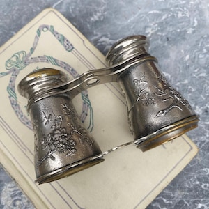 Pode incluir: Um par de óculos de ópera vintage com acabamento em prata metálica e padrões florais em relevo nos barris. Os binóculos possuem um mecanismo de ponte central e oculares circulares com bordas e detalhes visíveis em tom de latão.