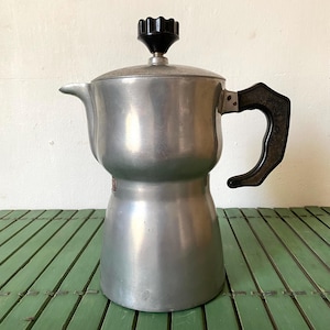 May include: A vintage silver stovetop espresso maker with a black handle and knob. The coffee pot has a classic design with a spout and a round base. The handle is curved for easy gripping. The espresso maker is made of metal and has a shiny finish.