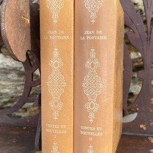 Peut inclure: Deux livres anciens de Jean de la Fontaine, intitulés "Contes et Nouvelles", en volumes I et II. Les livres ont une couverture orange clair avec des motifs et du texte blancs ornés. Ils sont exposés sur une étagère en bois.