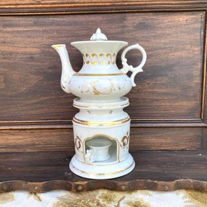Może przedstawiać: Biały porcelanowy podgrzewacz do herbaty ze złotymi akcentami. Podgrzewacz do herbaty ma górną część w kształcie czajniczka z pokrywką i uchwytem oraz podstawę z małą przegródką na podgrzewacz. Projekt obejmuje złote wykończenia i dekoracyjne wzory.