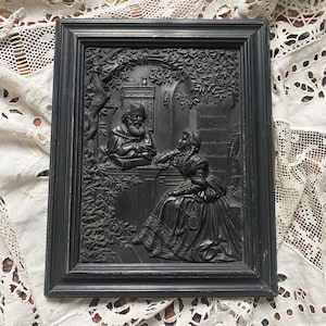 Op de afbeelding: Zwart omlijnde reliëfsculptuur met een man en een vrouw in een tuin. De man staat bij een raam, de vrouw zit. Het reliëf is gedetailleerd met bladeren en architectonische elementen.