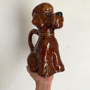 Puede incluir: Una jarra de cerámica marrón con forma de caniche y acabado brillante. La jarra tiene un asa y un pico en la nariz del perro. El perro está sentado erguido, con la cabeza en alto. Es un artículo decorativo.