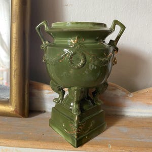May include: An olive green ceramic urn with two side handles, featuring gold-painted laurel wreath and swag detailing. The vessel is supported by three sculpted animal-paw feet that rest upon a square base with gold-painted decorative accents.