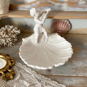 Könnte beinhalten: Eine weiße Keramikschale in Muschelform mit einem Cherub, der ein Saiteninstrument spielt. Die Schale hat einen gewellten Rand. Dekorativer Artikel.