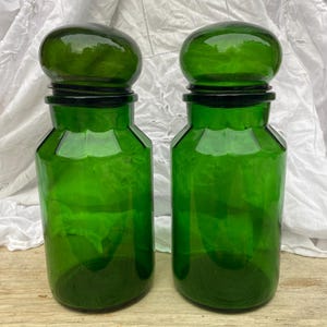Könnte beinhalten: Zwei passende Apothekergläser aus smaragdgrünem Glas mit abgerundeten, gewölbten Deckeln. Jedes Glas hat eine sechseckige Form mit glatten, flachen Seitenflächen und einem dicken, stabilen Boden. Das Glas ist durchscheinend und leuchtend grün.