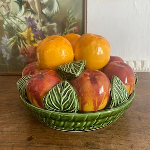 Puede incluir: Un frutero de cerámica lleno de fruta de cerámica naranja y roja. El cuenco es verde con un diseño de cesta tejida. La fruta de cerámica incluye naranjas y melocotones, con hojas verdes de cerámica.