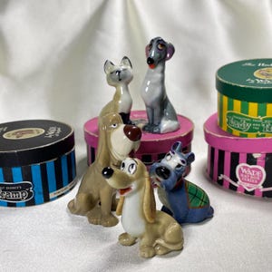 Puede incluir: Una colección de figuras de porcelana de perros y un gato, incluyendo personajes de la película de Disney "La Dama y el Vagabundo". Las figuras están en varias poses y colores, algunas sobre sombrereras decorativas.