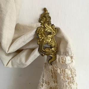 Peut inclure: Attache de rideau décoratif en métal doré avec un motif floral. L'attache est fixée à un rideau en tissu blanc.