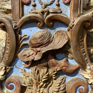 May include: Ornate, antique architectural detail with a hat and shoe motif. The relief sculpture features a hat and shoe in a light brown color, set against a light blue background. The surrounding frame is a darker brown with decorative scrollwork.