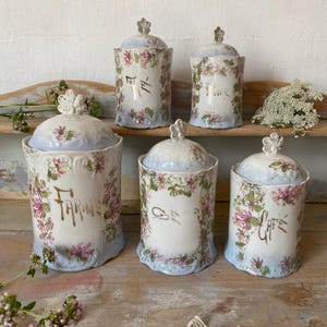 May include: A set of five ceramic canisters with lids, featuring a white base, light blue accents, and pink floral vine patterns. The canisters are labeled in gold script with the words FARINE, THE, POIVRE, SEL, and CAFE, each topped with a floral finial.