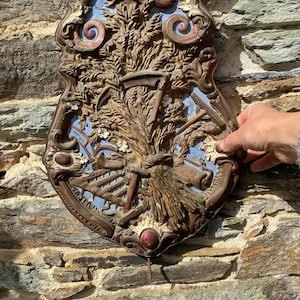 Peut inclure: Plaque murale antique en forme de bouclier avec un motif en relief détaillé. La plaque présente une gerbe de blé centrale, une faux et d'autres outils agricoles, avec un fond bleu. Le cadre est marron foncé avec des accents rouges et dorés.
