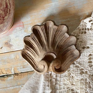 May include: A carved wooden decorative element shaped like a scallop shell with prominent radiating ridges and a central scroll detail at the base. The wood has a natural light brown finish with visible grain patterns and a smooth, contoured surface.