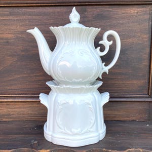 Op de afbeelding: Een witte porseleinen theepot met bijpassende warmer. De theepot heeft een gebogen tuit, een decoratieve handgreep en een deksel met een knop. De warmer heeft een geschulpte rand en een decoratief ontwerp. De set is een klassiek ontwerp.
