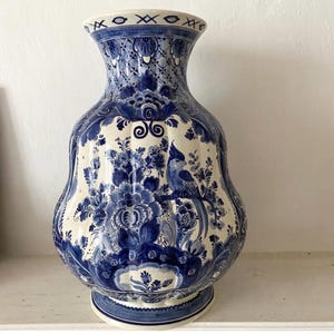 Può includere: Un grande vaso in ceramica con base bianca, decorato con complessi motivi floreali blu e l'illustrazione di un uccello. Il vaso presenta un corpo bulboso, un collo svasato e una base circolare, il tutto arricchito da dettagliati motivi blu.