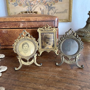 Peut inclure: Trois petits cadres photo en laiton orné avec des volutes complexes et des bases sur pied. Deux cadres sont circulaires avec des sommets décoratifs, tandis que le cadre central est rectangulaire. Un cadre circulaire contient un portrait vintage.