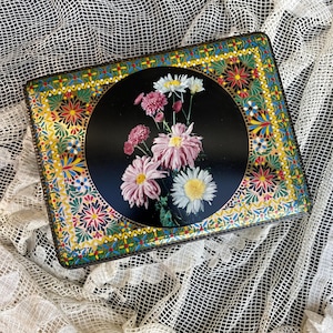 Könnte beinhalten: Rechteckige Vintage-Blechdose mit floralem Design. Die Mitte zeigt einen schwarzen Kreis mit rosa und weißen Chrysanthemen. Der Rand hat ein farbenfrohes, aufwendiges Muster mit gelben, grünen, roten und blauen Akzenten. Die Dose liegt auf einer weißen Spitzentischdecke.