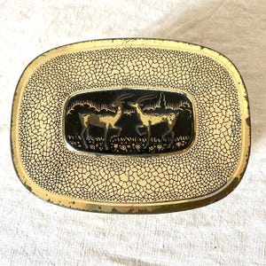 Op de afbeelding: Een vintage ovale blikken doos met een goud en zwart craquelé patroon. Het midden heeft een zwarte rechthoek met twee gouden herten silhouetten. De blikken doos heeft een gouden rand en een licht versleten uiterlijk.