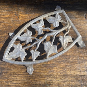 May include: Silver-coloured trivet featuring a leaf and vine design. The trivet is shaped like a quarter circle and has three small feet. The leaves and vines are intricately detailed, adding a touch of elegance to the kitchen accessory.