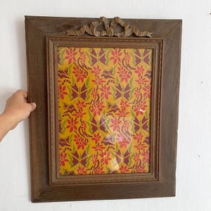 Può includere: Una cornice rettangolare in legno con un fiocco decorativo nella parte superiore. La cornice racchiude una stampa colorata con fiori rossi, foglie verdi e forme di farfalle viola su sfondo giallo.