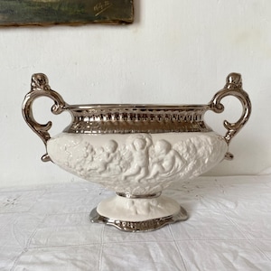 Könnte beinhalten: Eine weiße Keramikvase mit silberfarbenen Verzierungen und Griffen. Die Vase hat ein Reliefdesign von Engeln und einen Sockel. Der Rand und die Griffe haben eine metallisch silberne Oberfläche, die dem Dekorstück Eleganz verleiht.