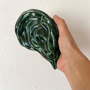 May include: A dark green ceramic sculpture with a glossy finish. The organic shape resembles a leaf or a shell, with textured, swirling details. The sculpture is held against a white background.
