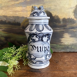 Può includere: Un vaso da farmacia in ceramica bianca e blu con coperchio, con la scritta "Pulpe". Il vaso presenta un motivo floreale decorativo ed è ambientato su uno sfondo in stile vintage.