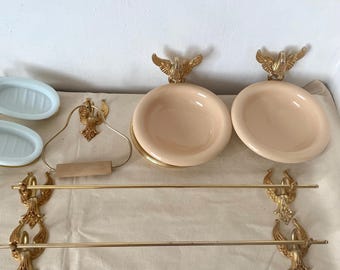 Puede incluir: Un conjunto de accesorios de baño vintage en dorado y crema. Incluye dos jaboneras, dos platos redondos con detalles de águila, un portarrollos de papel higiénico y un toallero. Los accesorios tienen un diseño clásico y ornamentado.