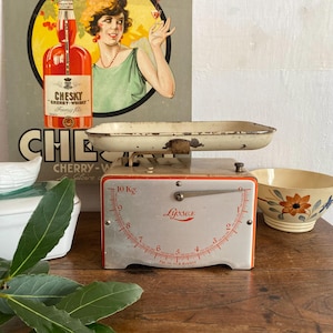 May include: Vintage scale with a cream-colored tray and a gray base with red accents. The scale reads up to 10 kg. A vintage advertisement for "Chesky Cherry-Whisky" is in the background.