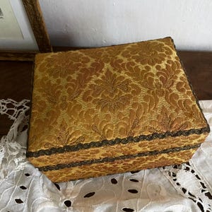 Op de afbeelding: Een rechthoekige, antieke sieradendoos met een gouden bloemenbrokaatpatroon. De doos is afgezet met donkerbruin kant en heeft een scharnierend deksel. De doos staat op een witte kanten doek.