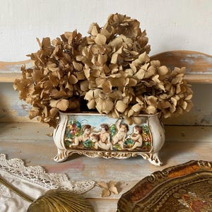 Peut inclure: Une jardinière rectangulaire en céramique à quatre pieds ornés, présentant un relief de quatre chérubins dans un jardin avec des touches de ciel bleu. La jardinière est remplie d'un grand bouquet d'hortensias séchés marron.