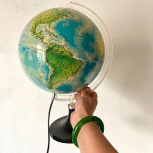 Peut inclure: Un globe terrestre vintage éclairé, avec un méridien en plastique transparent et une base noire. Le globe présente une carte détaillée avec des océans bleus et des masses terrestres vertes et jaunes. Un bracelet vert est visible.