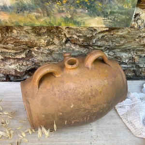 Può includere: Una grande brocca in terracotta marrone disposta orizzontalmente con una superficie ruvida e usurata. Il recipiente presenta due manici curvi sulla parte superiore, un'apertura circolare centrale e un piccolo beccuccio.