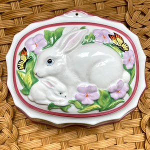 Peut inclure: Moule en céramique blanche en forme de lapin mère et de lapin bébé entouré de feuilles vertes et de fleurs violettes avec deux papillons jaunes et orange.
