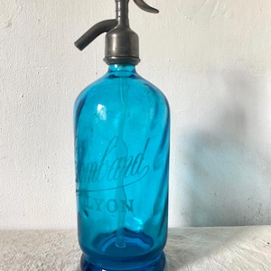 May include: Antique turquoise glass seltzer bottle with a metal dispenser. The bottle has a swirl design and the word "Lombard" and "LYON" are printed on the front. The dispenser is dark gray.