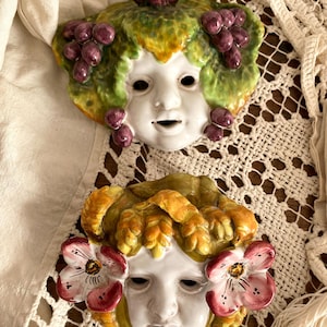 Peut inclure: Deux masques muraux en céramique avec des détails peints. Le masque du haut a un visage vert et jaune avec des raisins et des feuilles violets. Le masque du bas a un visage jaune et vert avec des fleurs roses.
