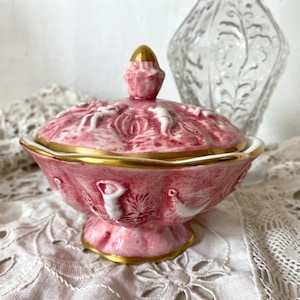 Może przedstawiać: Różowa ceramiczna miseczka na drobiazgi z pokrywką, ozdobiona złotymi detalami. Miseczka zdobiona jest wypukłymi wzorami kwiatowymi i wiciami. Pokrywka ma złoty guzik.