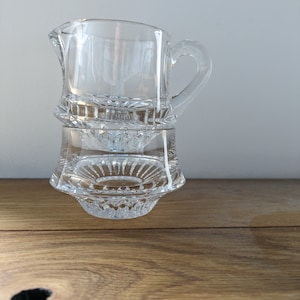 Può includere: Un'annaffiatoio in vetro trasparente con un design decorativo in vetro tagliato. L'annaffiatoio ha un manico e una base larga.