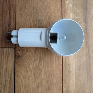 Könnte beinhalten: Eine weiße Strahlerlampe mit zylindrischem Sockel und einem breiten, schüsselförmigen Reflektor. Die Lampe verfügt über zwei Metallstifte am Ende für den elektrischen Anschluss und ein kleines, klares Glasfilament im schwarzen Sockel.
