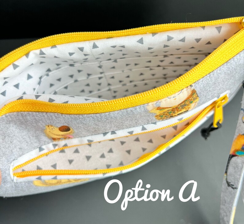 Puede incluir: Una bolsa gris con cremallera amarilla y forro estampado blanco y gris. La bolsa tiene dos compartimentos y presenta un peque&ntilde;o estampado de animales. El texto "Opci&oacute;n A" es visible en la imagen.