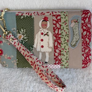 May include: A rectangular fabric wristlet pouch featuring a patchwork design of floral and holiday-themed patterns in red, green, and beige. The front displays a central applique of a figure in a white coat and red hat, plus a stocking ornament.