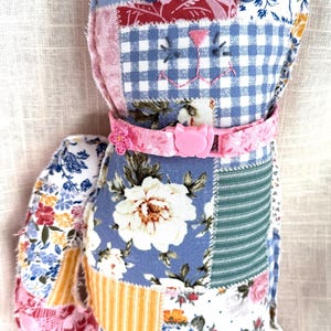 May include: A stuffed cat toy made from various fabric patches featuring floral, gingham, and striped patterns in blue, pink, white, and yellow. The cat has dark blue button eyes, an embroidered pink nose, and a pink plastic collar with a buckle.
