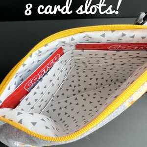 Puede incluir: Una bolsa con cremallera amarilla y gris con 8 ranuras para tarjetas. La bolsa est&aacute; abierta y el interior est&aacute; forrado con una tela gris con peque&ntilde;os tri&aacute;ngulos blancos. Dos tarjetas rojas con la palabra "Costco" son visibles en las ranuras.