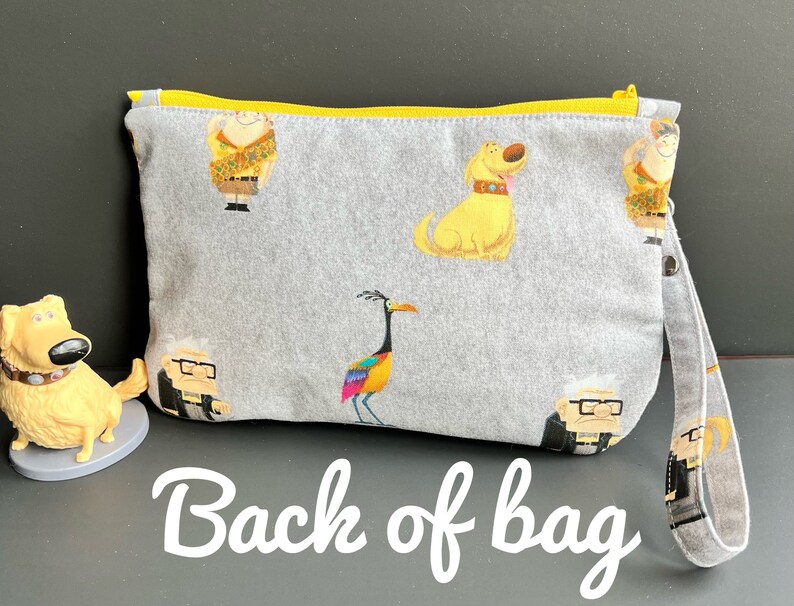 Puede incluir: Bolsa de cremallera gris con una cremallera amarilla y una correa amarilla. La bolsa tiene una variedad de personajes de dibujos animados coloridos impresos en ella, incluyendo un perro, un p&aacute;jaro y un hombre con gafas. El texto "Back of bag" est&aacute; escrito en blanco sobre un fondo gris debajo de la bolsa.