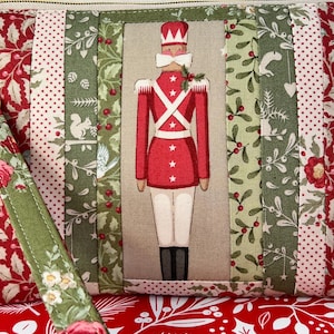 May include: A festive Christmas-themed pouch with a red and green color scheme. The central design features a toy soldier in a red uniform with white accents. The pouch is made of fabric with various patterns, including floral and dotted designs.