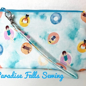 May include: A light blue zippered pouch with a wrist strap. The pouch has a watercolor design of people and animals floating in inner tubes. The text "Paradise Falls Sewing" is printed on the bottom of the image.